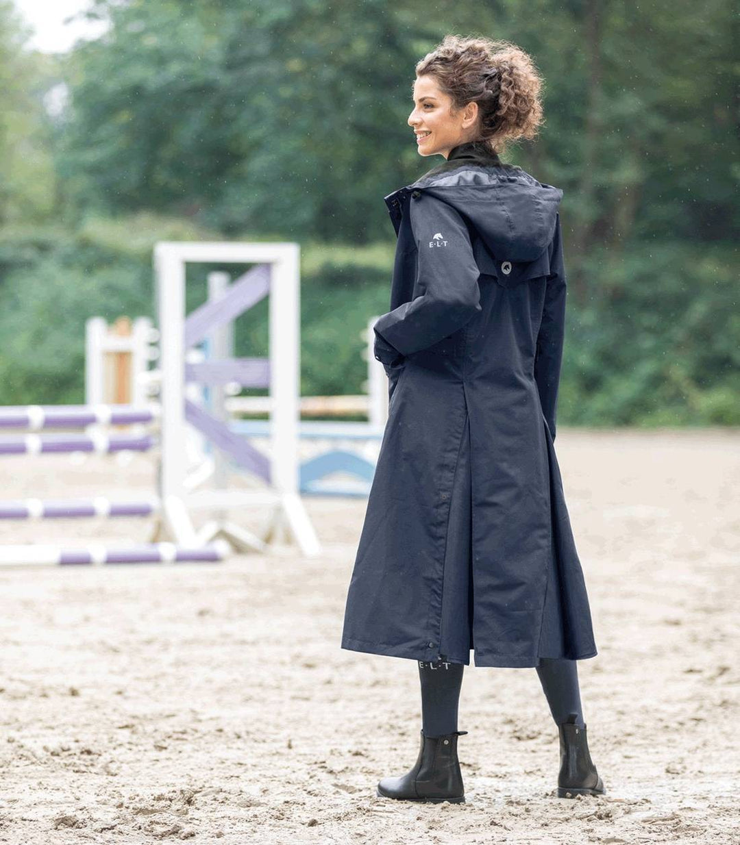Woman wearing the ELT Fehmarn Long Riding Raincoat in a stable setting, highlighting its long length and weather-resistant features.