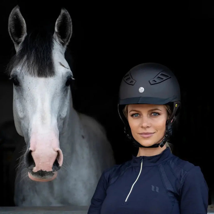 Rider wearing the Back On Track EQ3 Lynx Smooth Riding Helmet with MIPS, standing beside a white horse, showcasing its stylish design.
