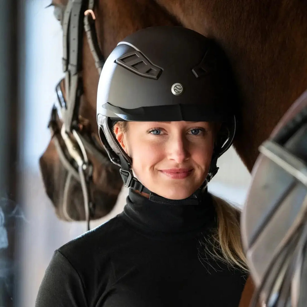 Rider wearing the Back On Track EQ3 Lynx Smooth Riding Helmet with MIPS, standing beside a horse, showcasing its stylish and protective design.