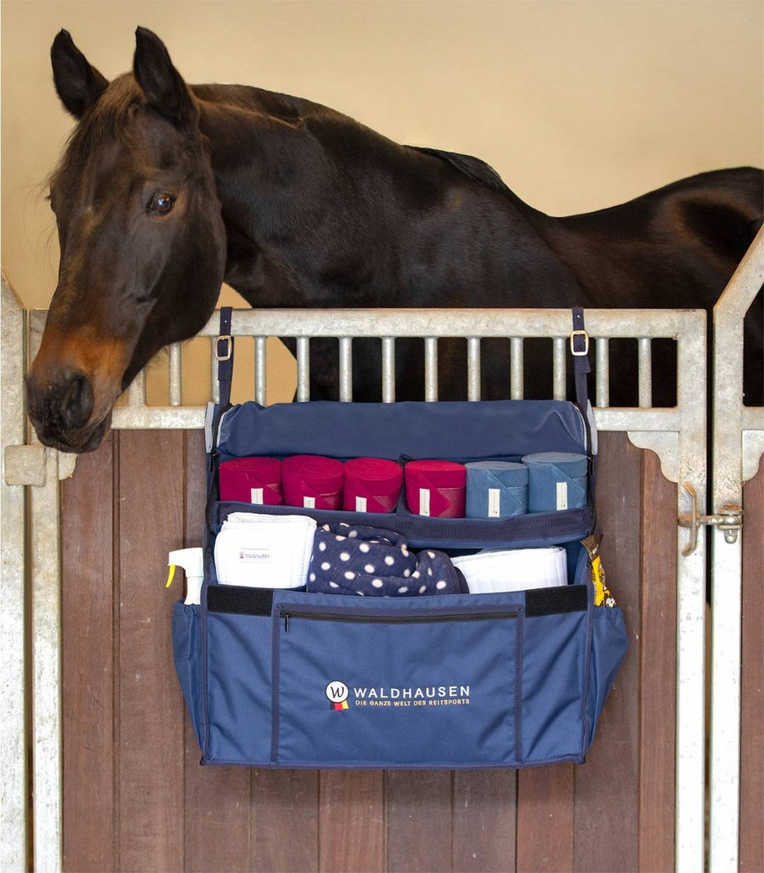 Waldhausen Stable Organiser Bag hanging on a stable door, filled with equestrian essentials like bandages and grooming tools, next to a horse.