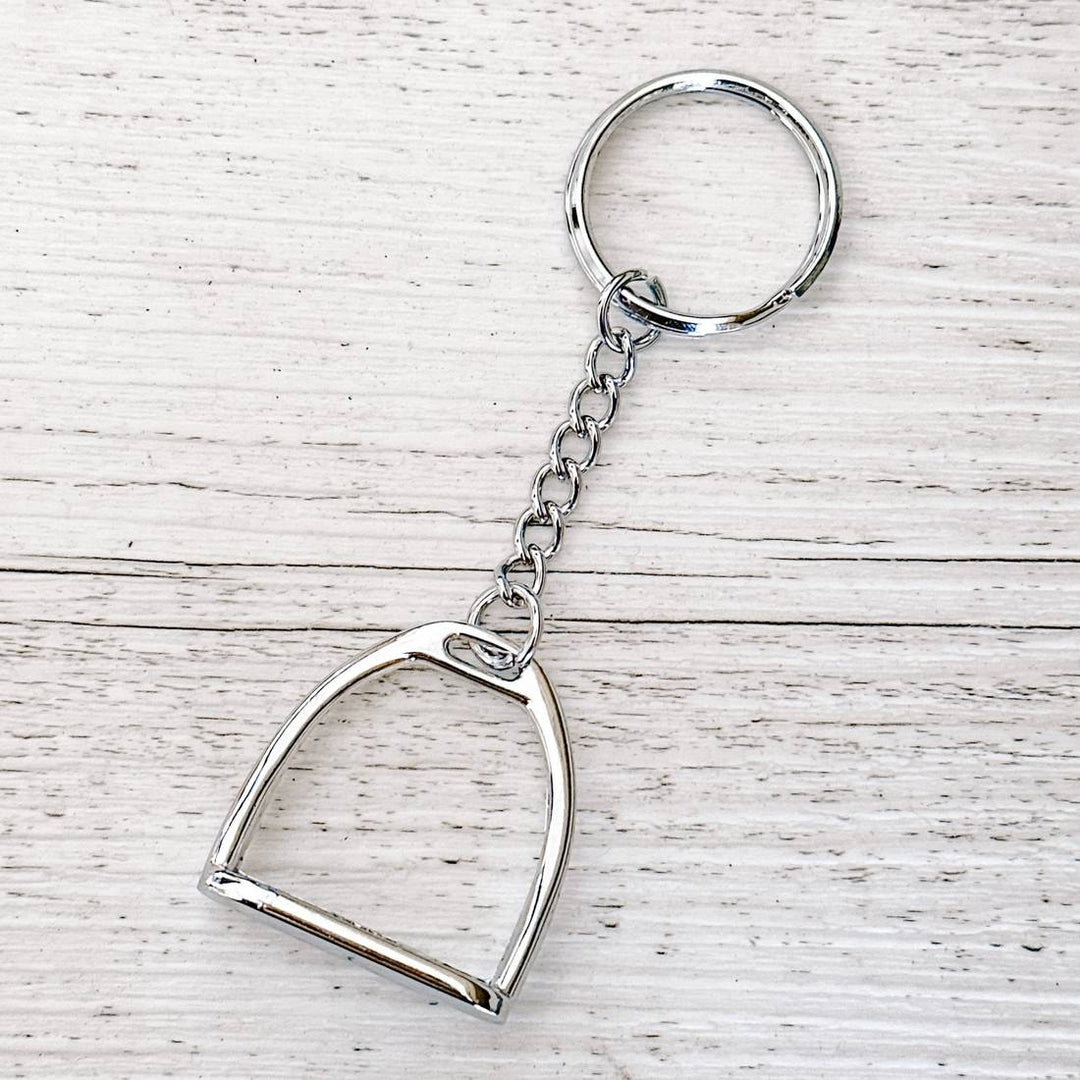 Equestrian-themed silver stirrup keychain displayed on a wooden surface, featuring a stirrup charm and chain.