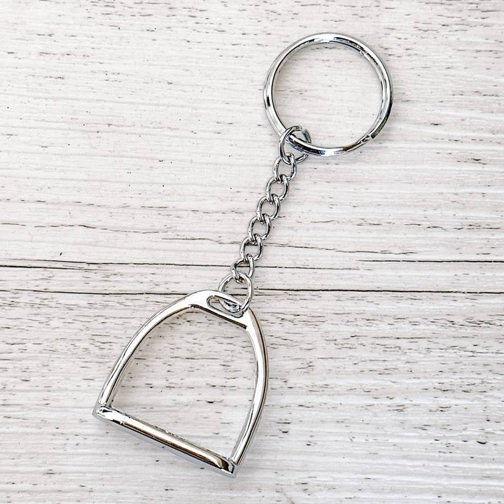 Equestrian-themed silver stirrup keychain displayed on a wooden surface, featuring a stirrup charm and chain.