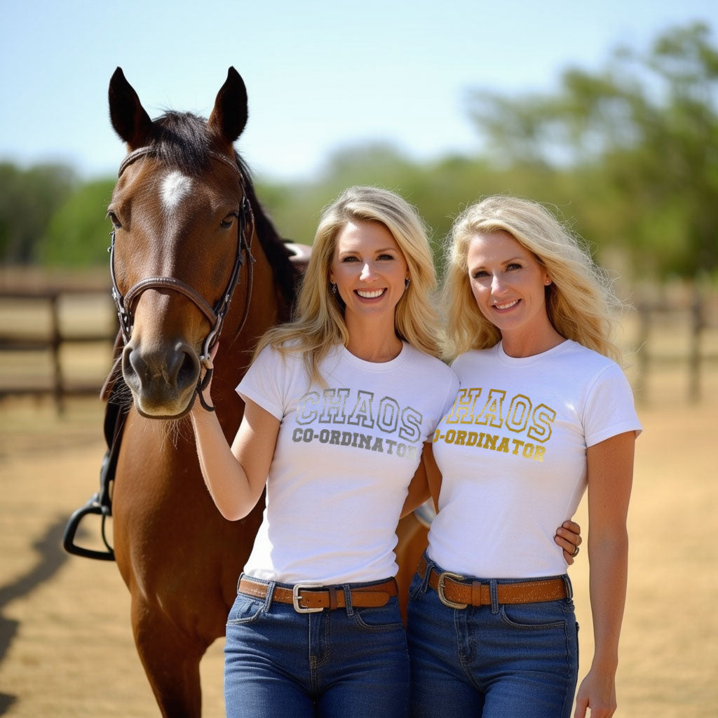 Two women wearing Chaos Co-Ordinator Tees in white with gold lettering, posing with a horse in an outdoor equestrian setting.