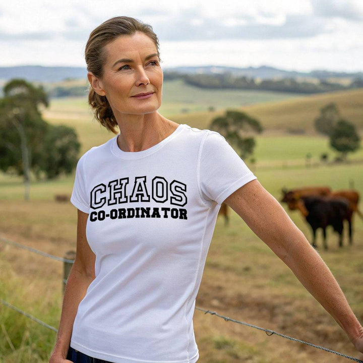 Woman wearing a white Chaos Co-Ordinator Tee with black lettering, standing in a scenic rural landscape with cattle in the background.