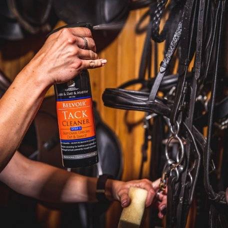 Person using Belvoir Step 1 Tack Cleaner Spray on leather bridles in a stable setting, showcasing its effectiveness in cleaning tack.