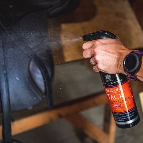 Hand spraying Belvoir Step 2 Tack Conditioner Spray onto a black leather saddle, showcasing its ease of use for leather care.