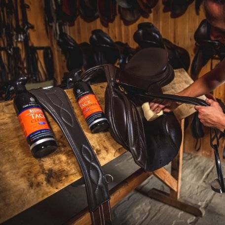 Belvoir Step 2 Tack Conditioner Spray bottles on a wooden table next to leather saddles, demonstrating its use in equestrian tack maintenance.